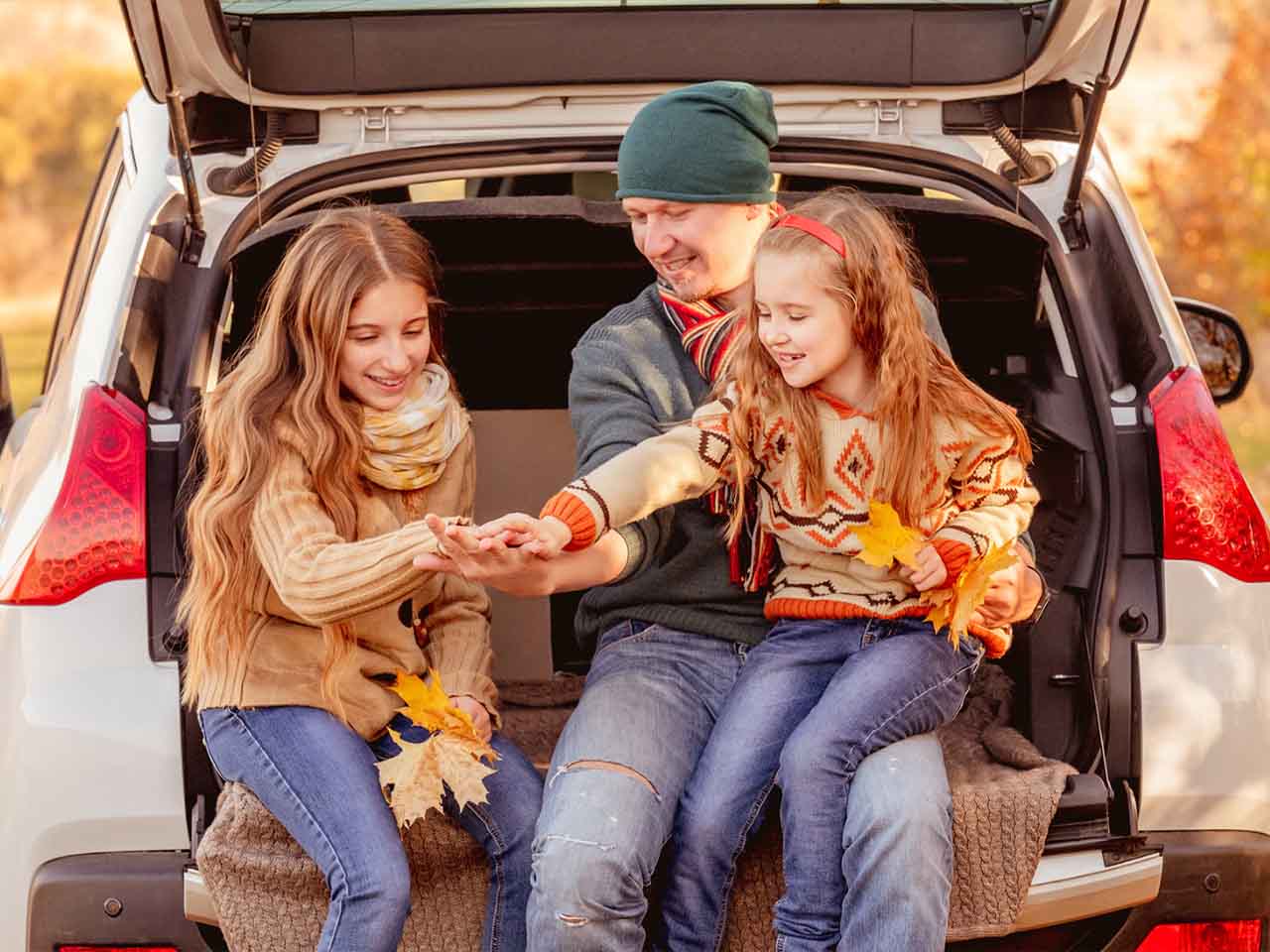 Father and two daughters sitting in the open trunk of a car, smiling and holding autumn leaves outdoors.