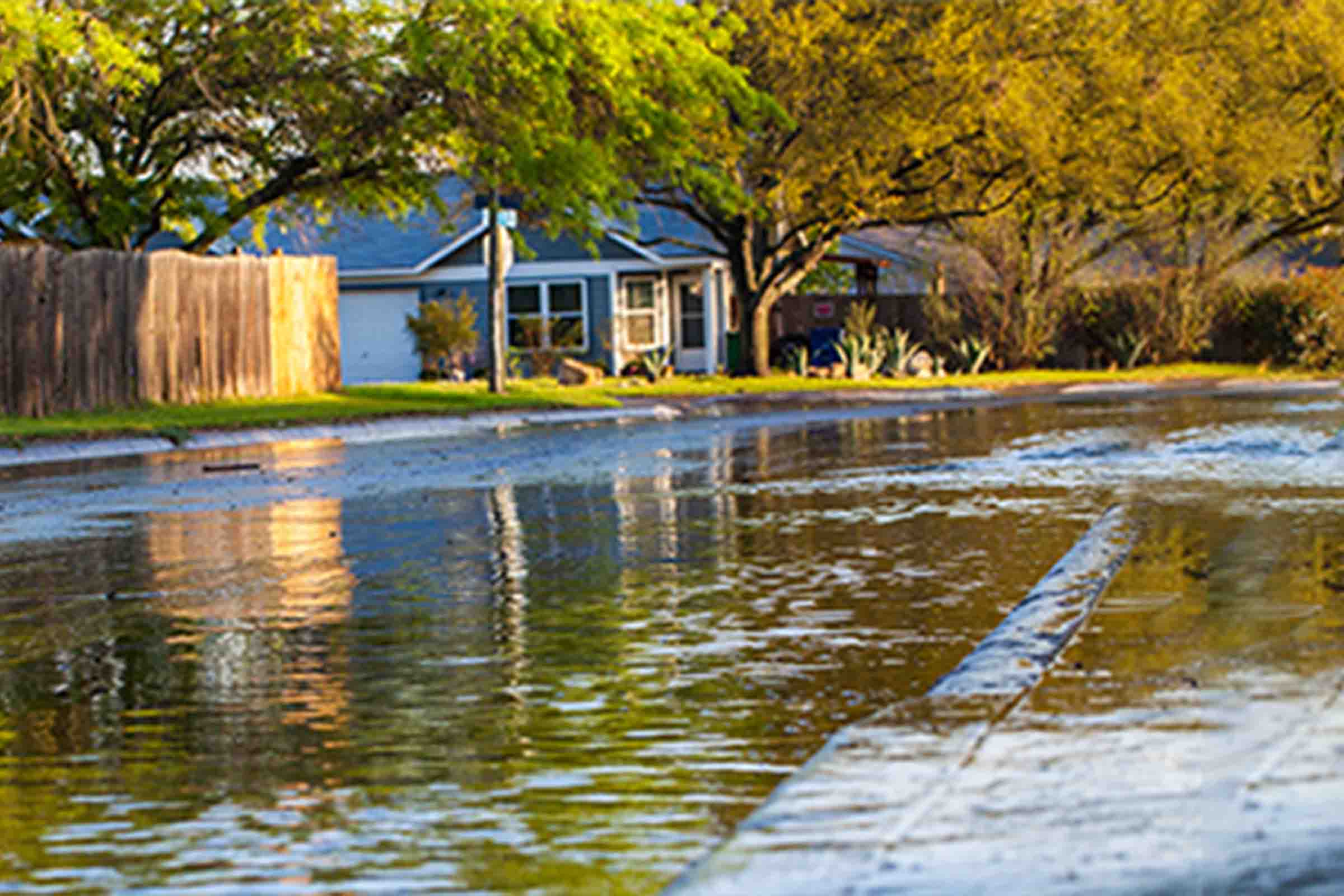 A tree-lined residential street with a small blue house beside a flooded roadway reflecting the autumn-colored trees.