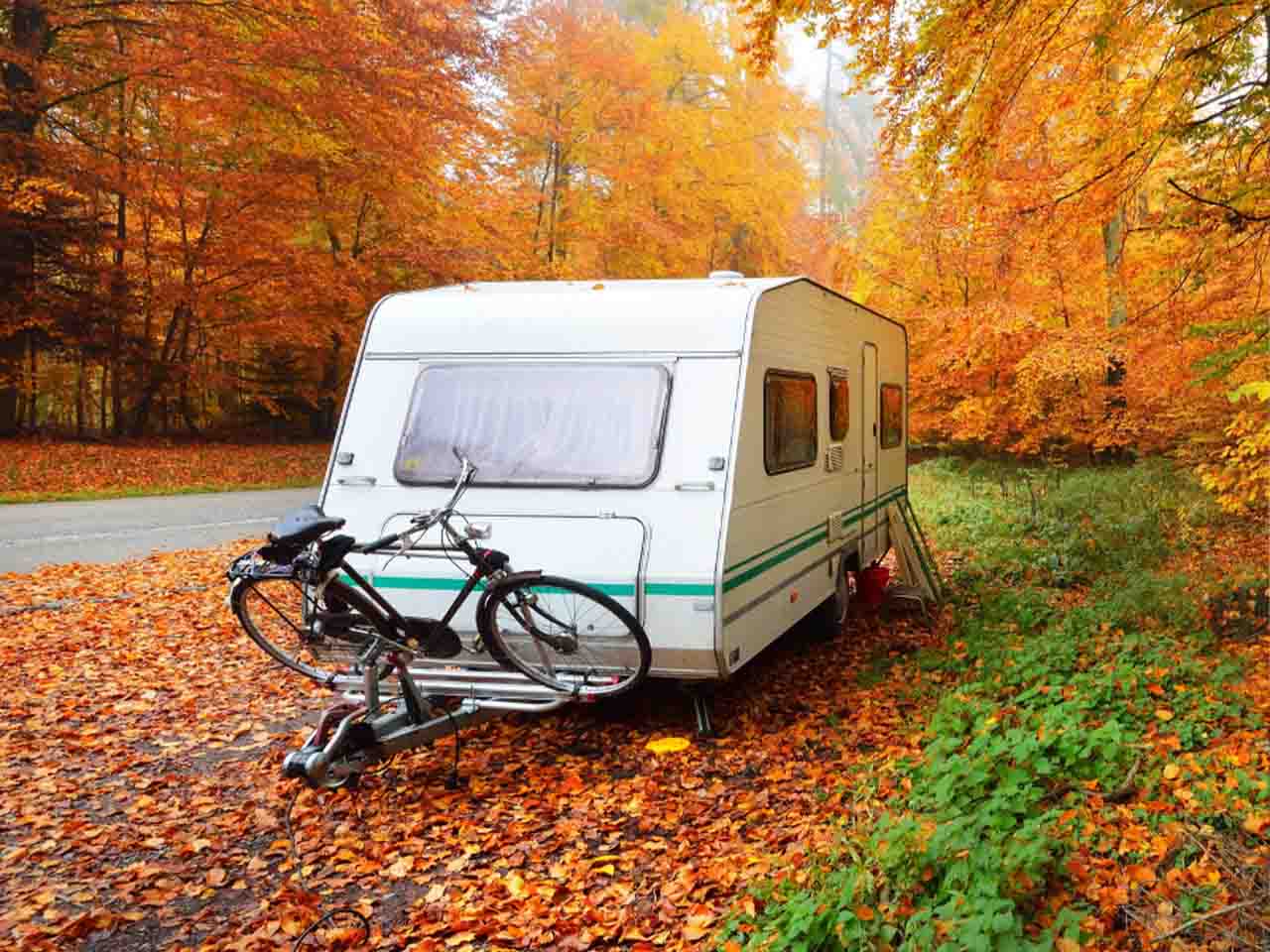 White camper trailer with two bicycles parked beside a forest road amid vibrant autumn foliage.