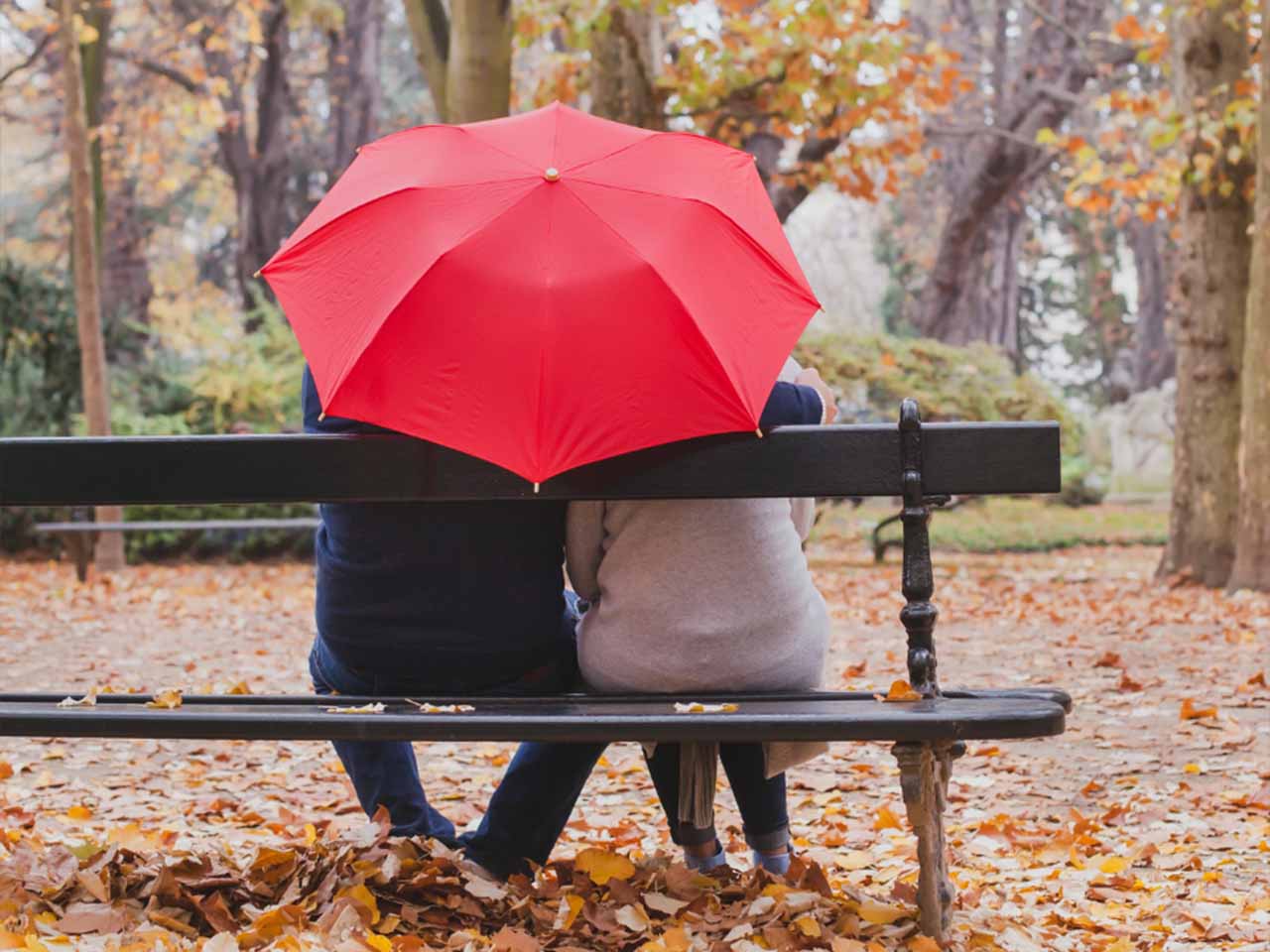 Two people sit together on a park bench beneath a red umbrella surrounded by fallen autumn leaves.